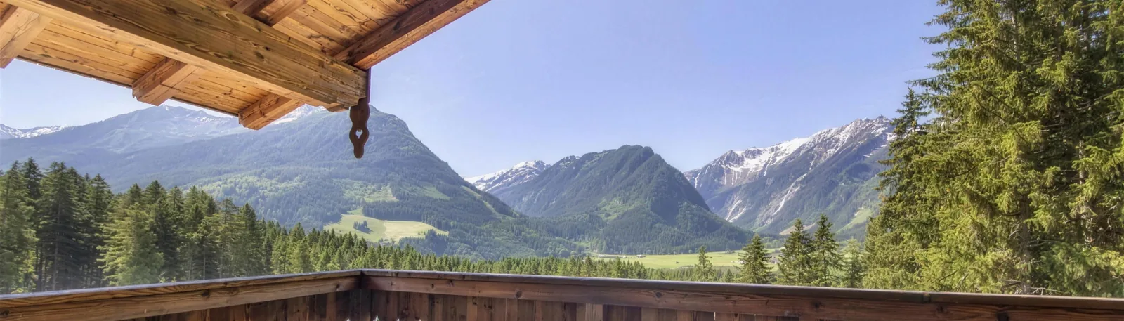 Blick von der Terrasse des Chalets über das Tal auf die Berge, oben der Dachbalken, unten das Holzgeländer