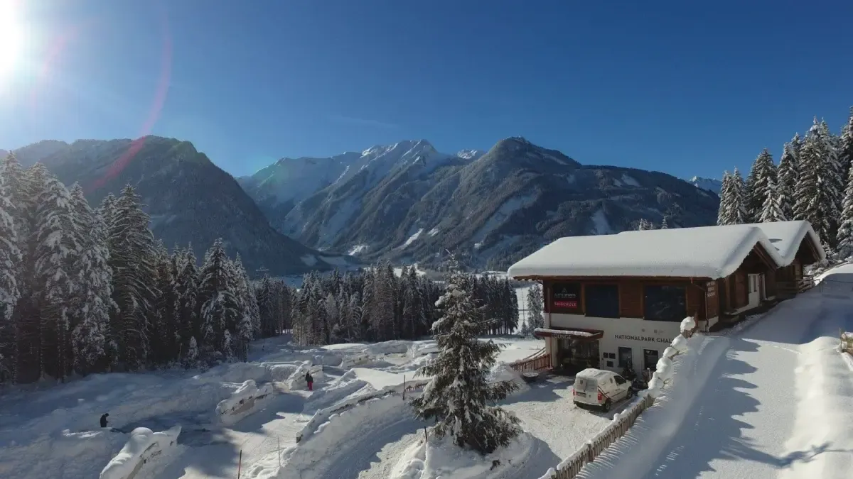Verschneite Fichten, tiefstehende Sonne, das Gebäude am Hang und dahinter das Tal mit der Gipfelkette