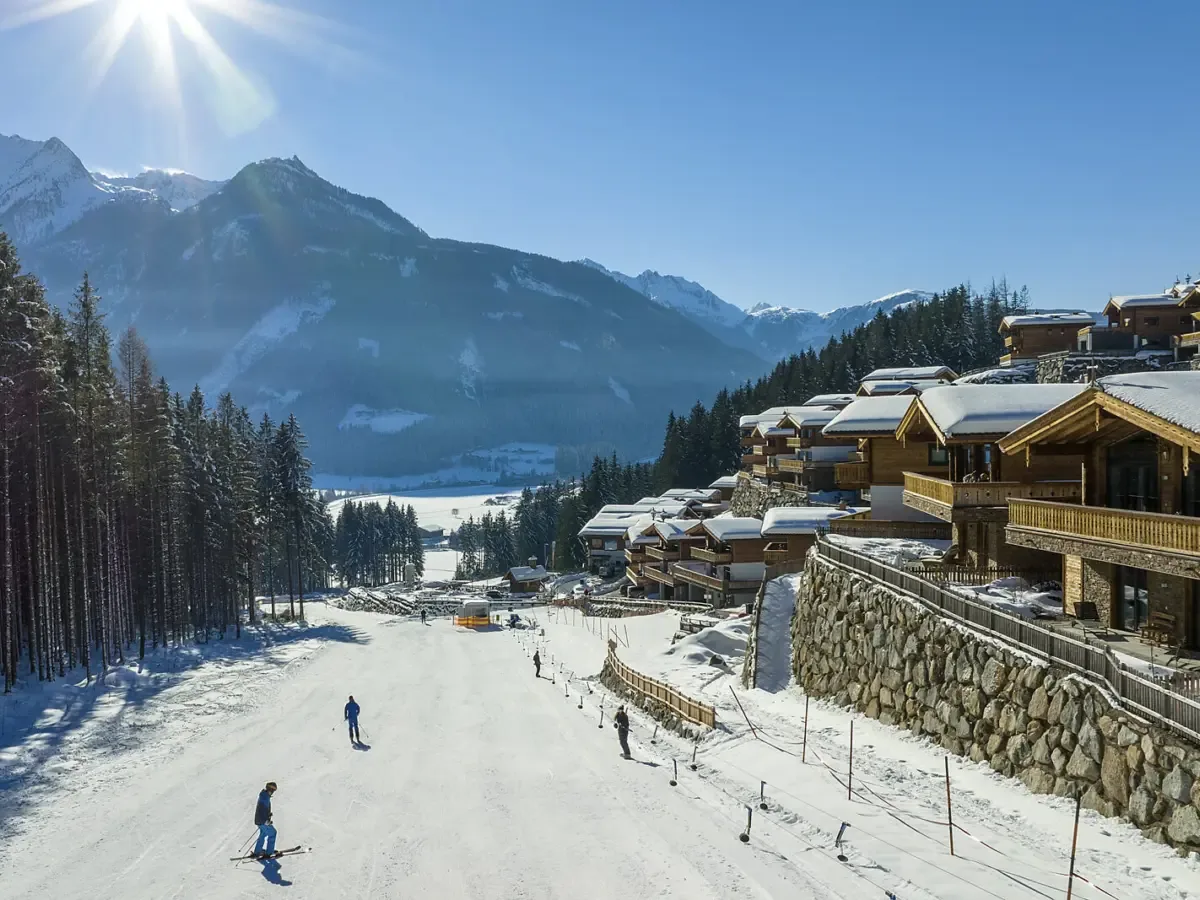 Die Piste am Chaletdorf mit Skifahrern, dahinter das Tal und die Gipfel