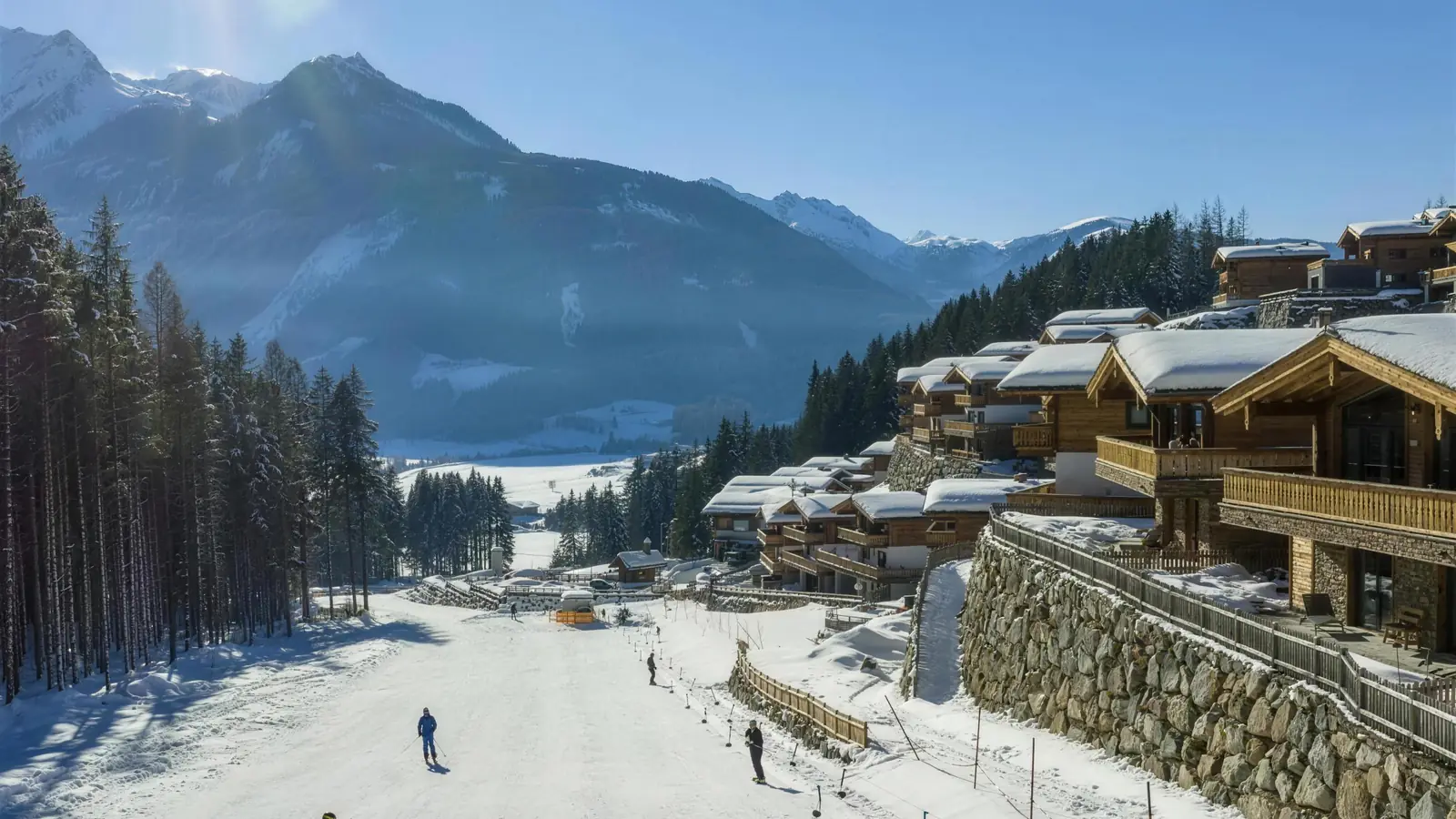 Die Piste am Chaletdorf, Skifahrer darauf, dahinter das Tal und die Gipfel der Wildkogel-Arena