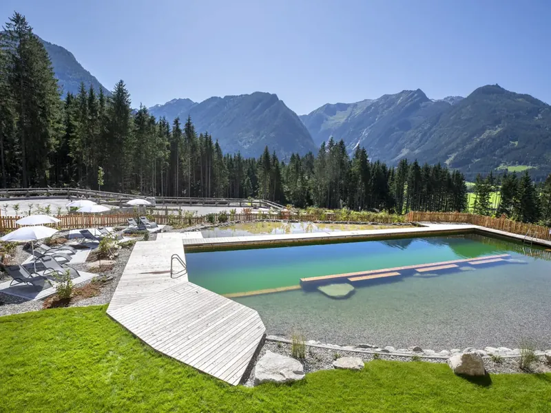 Der Naturpool des Chaletdorfs mit Holzsteg, dahinter Wald und Berge