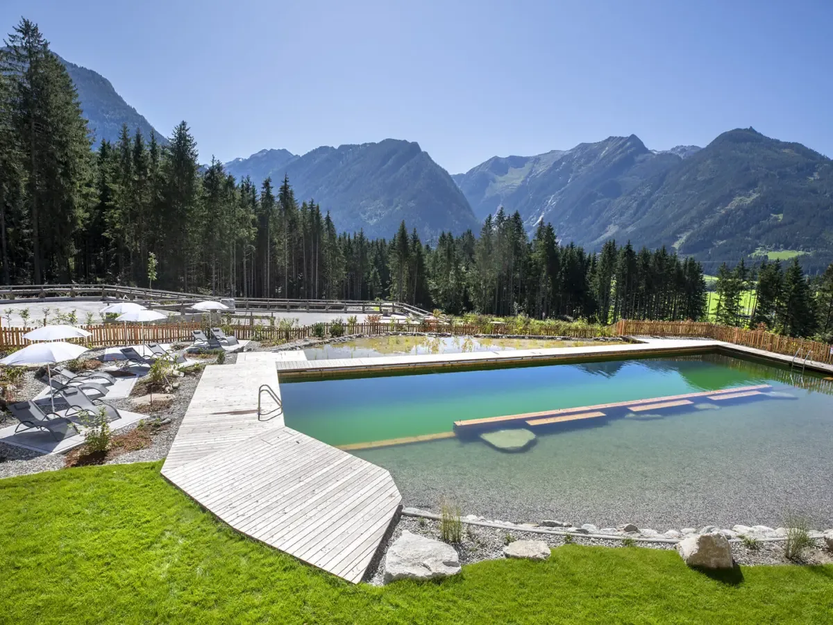 Der Naturpool im Chaletdorf, türkisfarbenes Wasser, Holzsteg mit Liegen, dahinter Wald und Berge