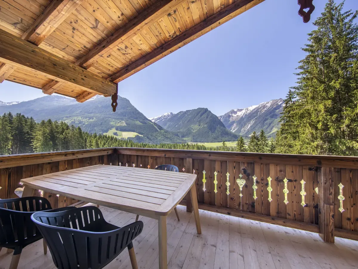 Die überdachte Terrasse mit Tisch und Stühlen, davor Fichten und das grüne Tal mit den Bergen
